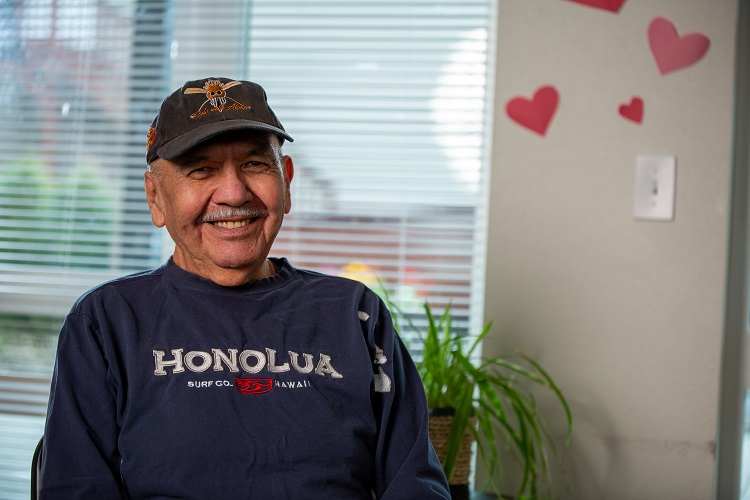 Curtis, wearing a cap and a blue long-sleeve shirt, smiles at the camera