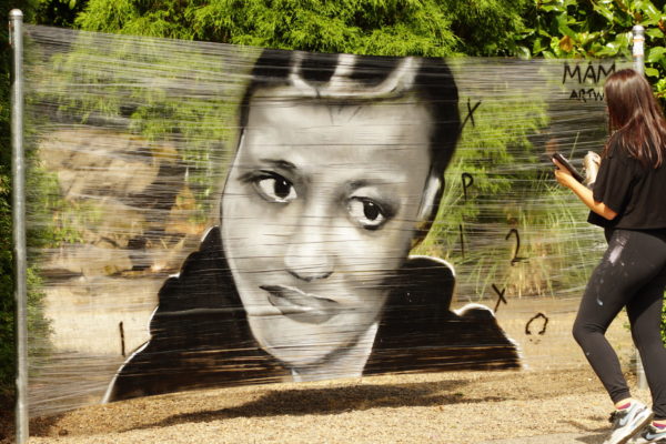 artist spray painting mural on stretched out cellophane; artist has long brown hair and wears all black. The art piece features a Black femme wearing a black hoodie.