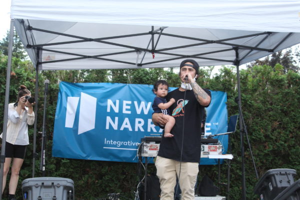 Talilo holding his baby talking on the mic onstage, the New Narrative banner behind him.