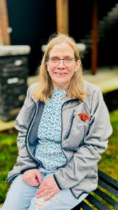 Photo of Jackie, seated, and looking at the camera. She has shoulder-length blonde hair and wears a grey Cardinals sweater over a blue outfit.