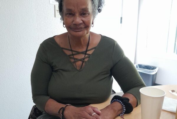 Image of Velda sitting at a table, hands touching on the table in front of her. She is wearing a dark green top and a headband, and smiling.