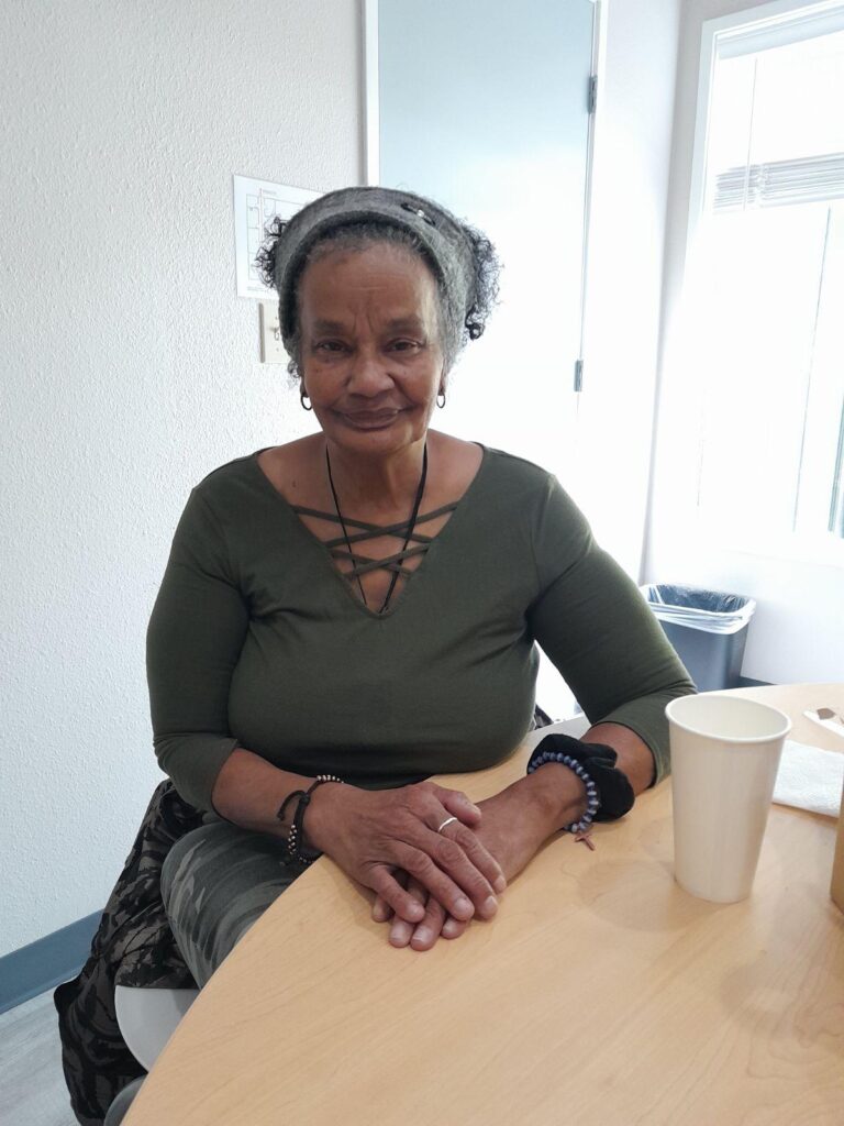 Image of Velda sitting at a table, hands touching on the table in front of her. She is wearing a dark green top and a headband, and smiling.