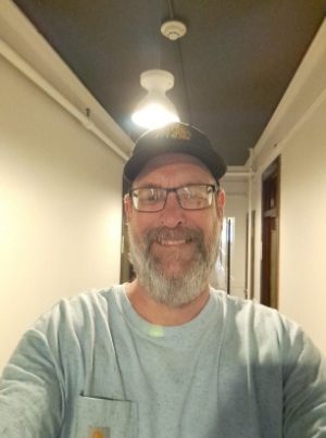 William, a white-presenting masculine person with a beard and glasses and a baseball cap, smiles at the camera